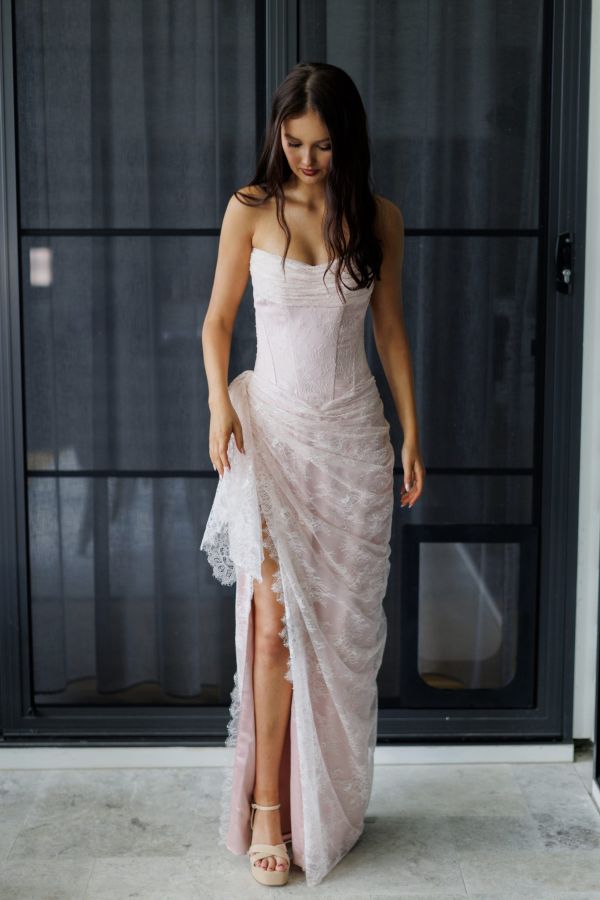 Model wearing the blush Bella Gown by Gossip Gowns, featuring a strapless lace corset bodice, draped floor-length skirt and high leg split.