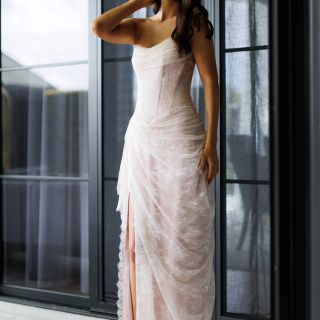 Model wearing the blush Bella Gown by Gossip Gowns, featuring a strapless lace corset bodice, draped floor-length skirt and high leg split.