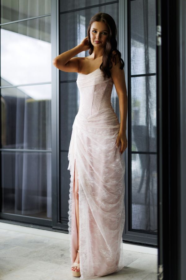 Model wearing the blush Bella Gown by Gossip Gowns, featuring a strapless lace corset bodice, draped floor-length skirt and high leg split.
